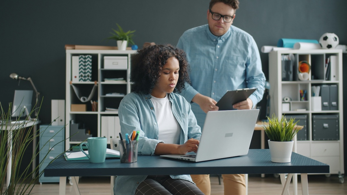A Black woman with curly hair works on a laptop at an office desk. Behind her, a white man wearing glasses is holding a clipboard and looking at her screen. It is a modern office environment with shelves, office supplies, and decorative plants.