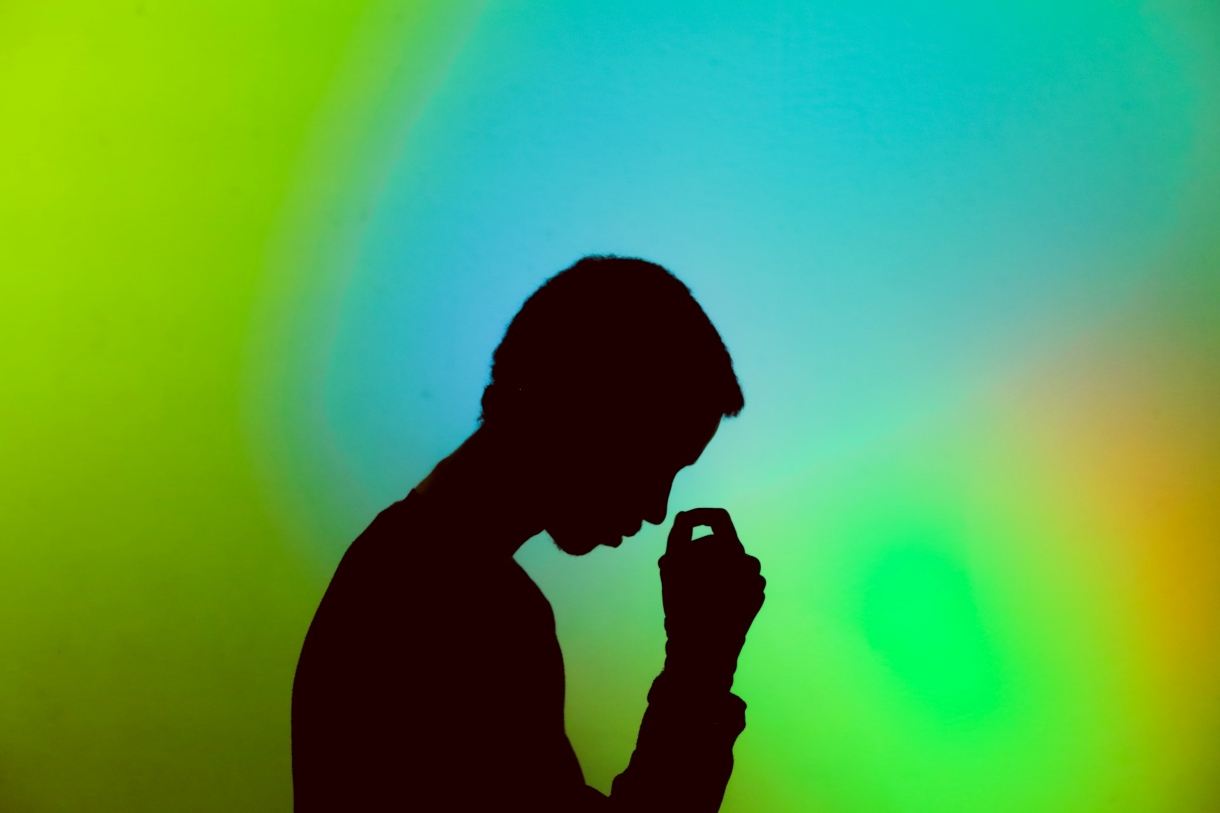 Silhouette of a person with their head tilted forward and their hand held near their face, suggesting worry or introspection. In the background, a gradient of vibrant green, blue, and yellow tones creates a contrast with the dark figure