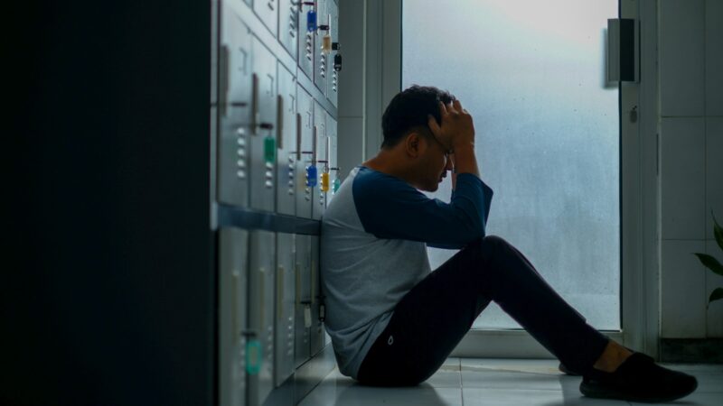 A man sitting on the floor in a hallway, leaning against cabinets with his head resting on his hand, in a position suggesting exhaustion or distress. A cold light shining through the doorway in the background reinforces the feeling of isolation and emotional tension.