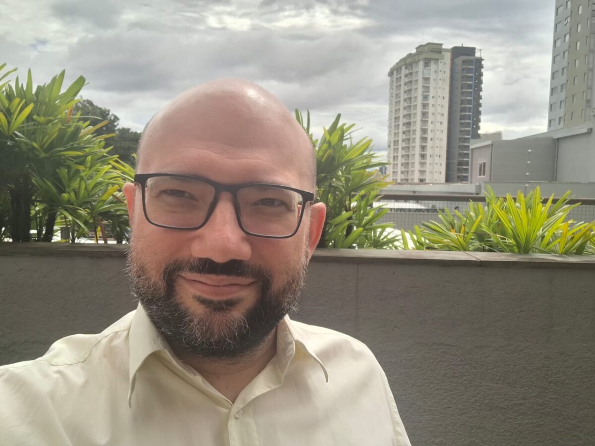 A bald man with glasses and a beard smiles in an outdoor selfie, wearing a white shirt. A modern building and trees are in the background.