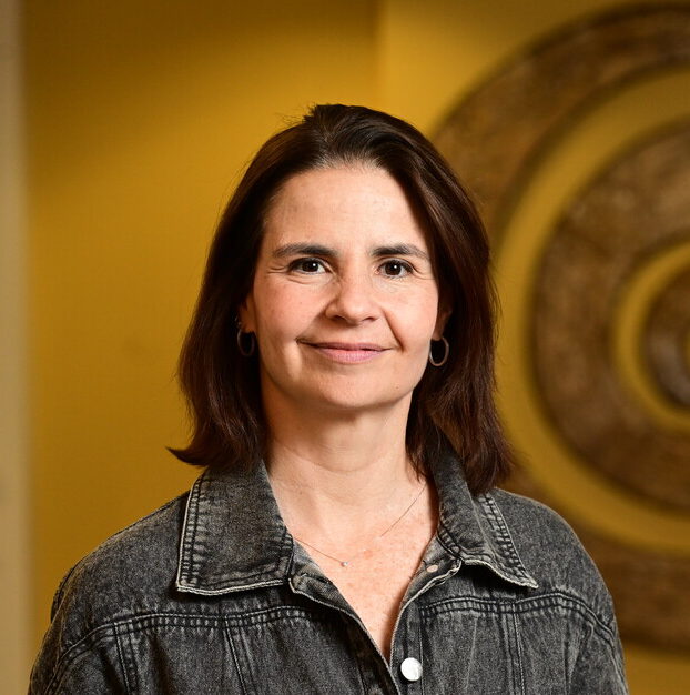 Retrato da médica infectologista Marina Caskey, da Universidade Rockefeller, em ambiente interno iluminado com tom amarelado. Ela usa uma jaqueta jeans e sorri levemente para a câmera. Ao fundo, uma escultura circular metálica em espiral compõe o cenário.