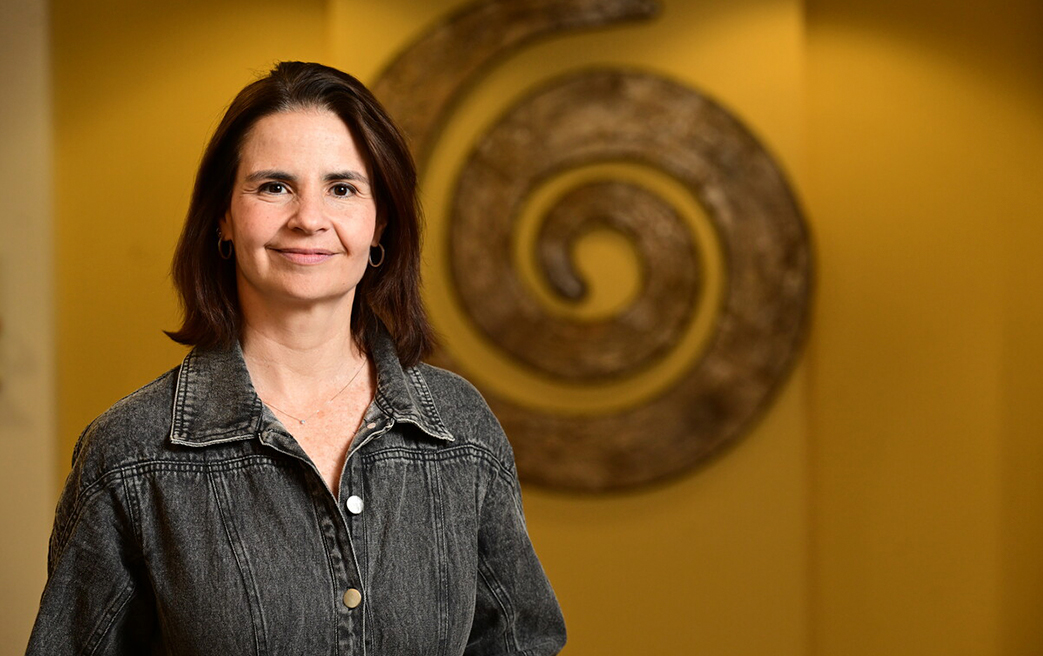 Portrait of infectious disease physician Marina Caskey, of Rockefeller University, indoors with warm amber lighting. She is wearing a denim jacket and smiling at the camera. A circular metallic sculpture appears in the background.