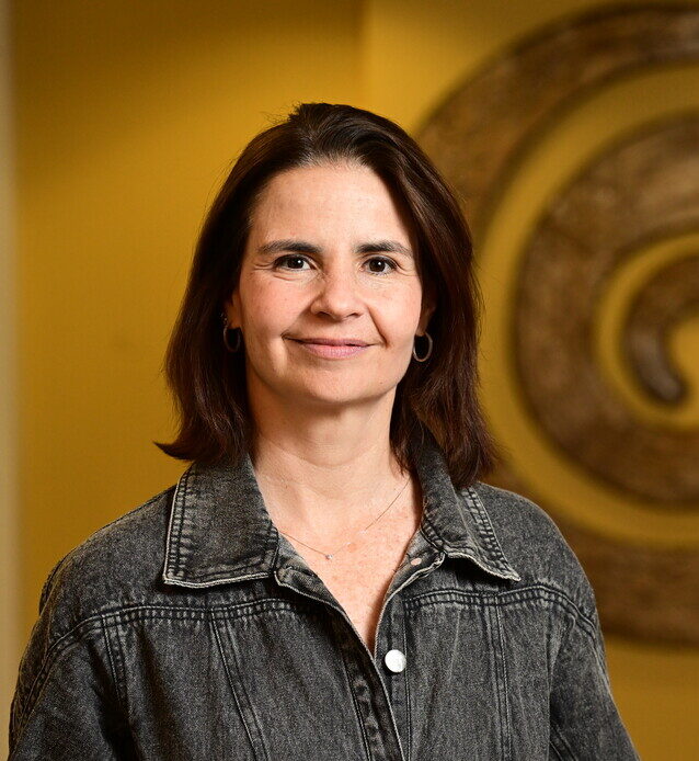 Portrait of infectious disease physician Marina Caskey, of Rockefeller University, indoors with warm amber lighting. She is wearing a denim jacket and smiling at the camera. A circular metallic sculpture appears in the background.