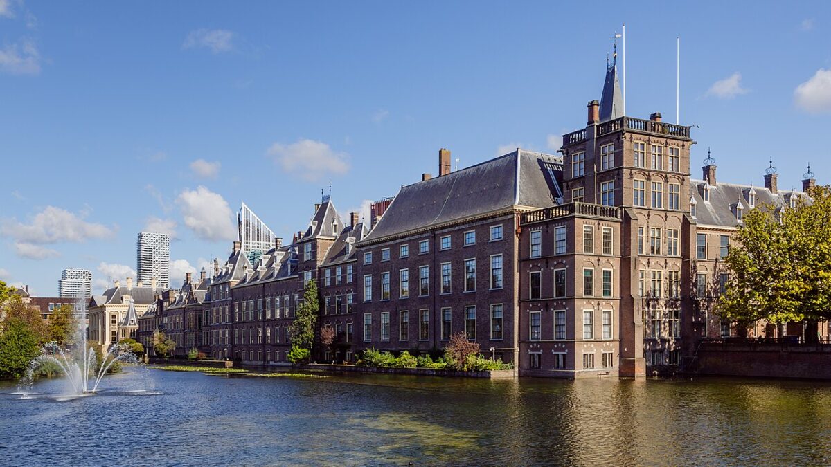 Photograph of the Binnenhof complex, home to the Dutch parliament, in The Hague. The historic architectural ensemble of dark bricks and Dutch Gothic-style roofs is seen on the shores of Lake Hofvijver, with a fountain on the water.