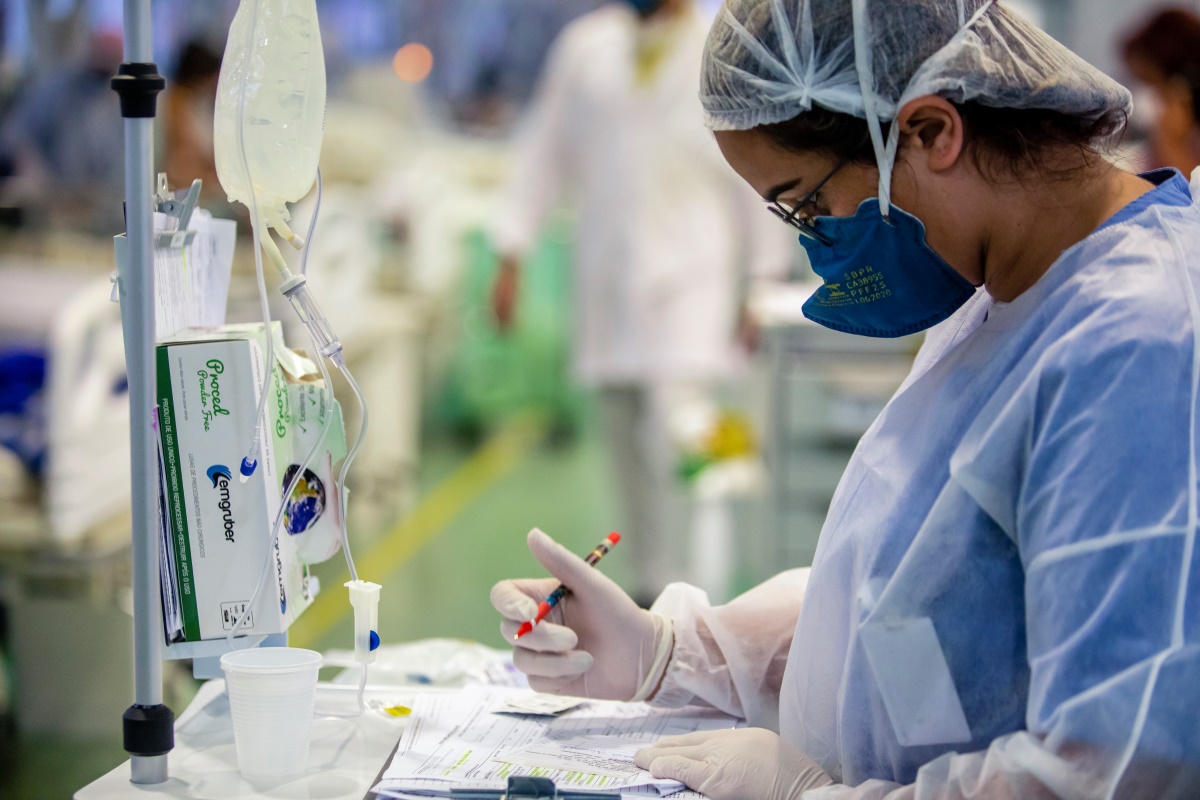 A healthcare professional in a hospital setting records information in a patient’s chart while monitoring equipment beside a patient in an intensive care unit (ICU). Wearing a mask, cap, and gloves, they work among medical devices and clinical support systems.