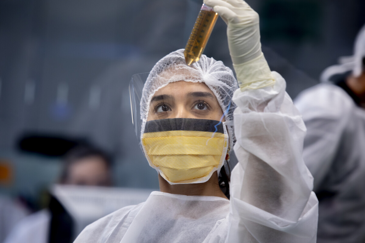 Pesquisadora, com equipamentos de proteção individual, observa atentamente um tubo de ensaio com líquido amarelado em ambiente de laboratório
