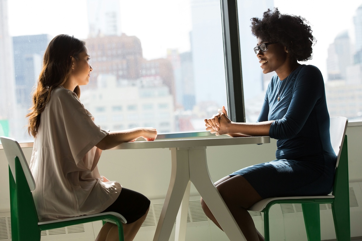 Two women sitting face-to-face at a table in a corporate environment. In the background, large windows with a view of urban buildings. The Black woman on the right, with afro hair and glasses, is smiling. The white woman on the left, with long straight hair, watches her attentively.