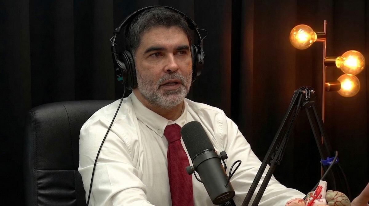 Middle-aged white man with dark graying hair and a short beard, wearing a white shirt and burgundy tie. He sits in a black chair, with black over-ear headphones, speaking into a studio microphone. The background is dark, illuminated by warm copper lamps. On the right side of the image, part of an anatomical brain model is visible.