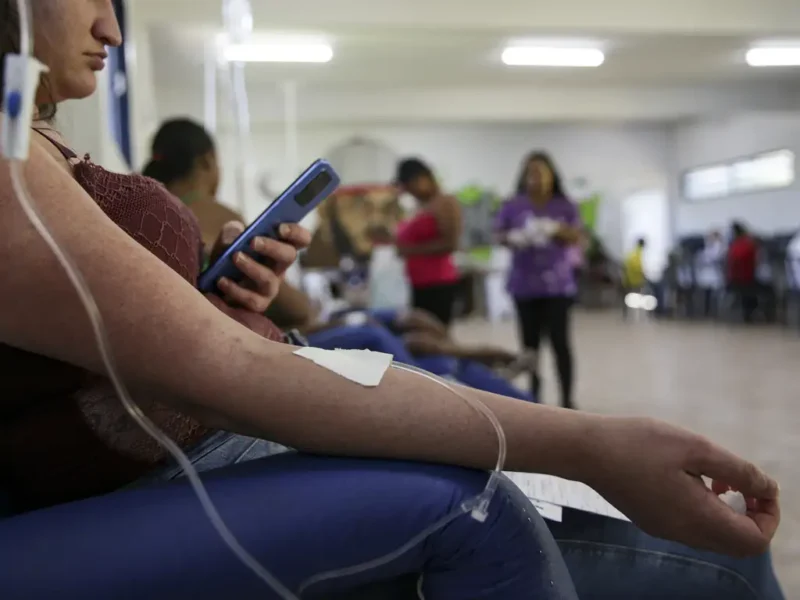A woman wearing a burgundy blouse receives care with an intravenous line in her right arm at a health center. She is holding a blue cellphone in her left hand. In the background, other people are seated receiving care, and a professional in a purple blouse is walking around the large, well-lit space