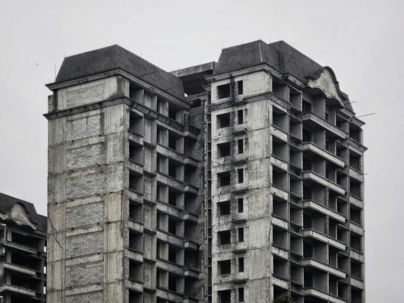 Vista em close de três edifícios residenciais de alto padrão abandonados, fotografados em preto e branco sob céu encoberto. As torres de concreto aparente, com cerca de quinze pavimentos, apresentam aberturas sem esquadrias, fachadas sem revestimento e fiação exposta.