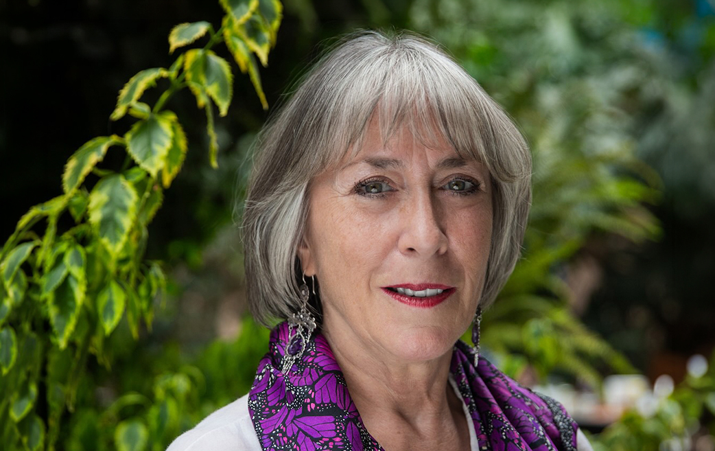 Photograph of Gabriela Dutrénit, a woman with short gray hair, wearing dangling earrings, a purple and black silk scarf, and a white top. She bears a half-smile, standing in front of an out-of-focus plant-filled background.