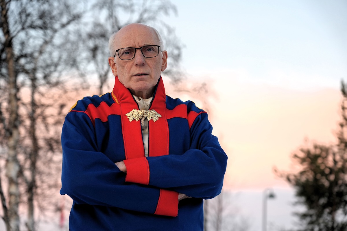 A white man with gray hair and black-framed glasses, appearing between 65 and 75 years old, stands with arms crossed and a serious expression. He wears a blue traditional Sami outfit with red trim and a gold brooch. Behind him are leafless trees and a pink-toned Arctic sunset sky.