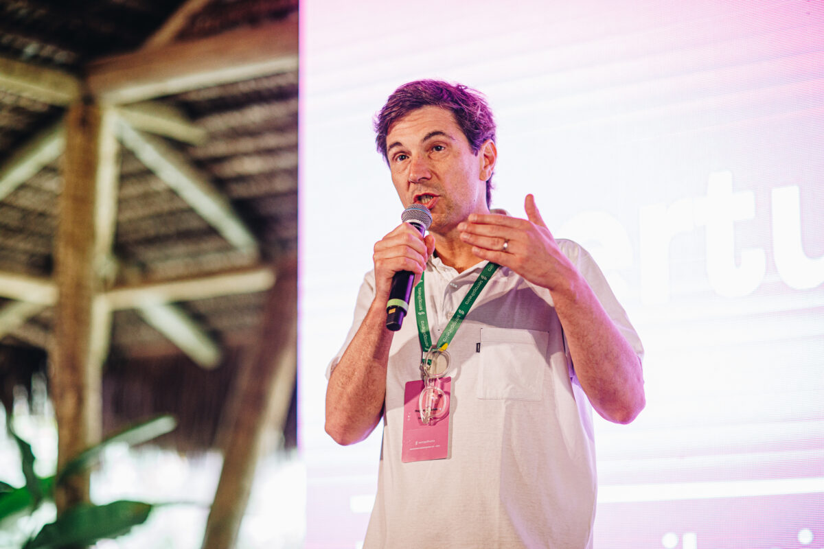 Homem branco de meia-idade, cabelos castanhos, vestindo camisa branca e crachá com cordão verde, segura microfone e gesticula com a mão direita durante apresentação. Ao fundo, telão com iluminação rosada e estrutura de palco com teto de palha.
