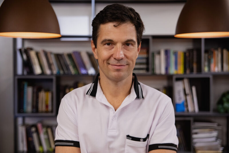 A middle-aged man with short brown hair, a calm expression, and a slight smile, wearing a white polo shirt with black detailing. In the background, there is a bookshelf and lights hanging from an arched ceiling.