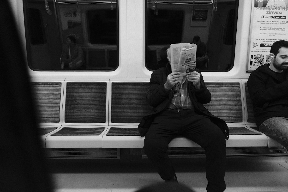 Black-and-white photograph of an elderly man sitting in a subway car, reading a newspaper that covers his face. In the background, other passengers appear out of focus in their seats. It is an everyday urban setting.
