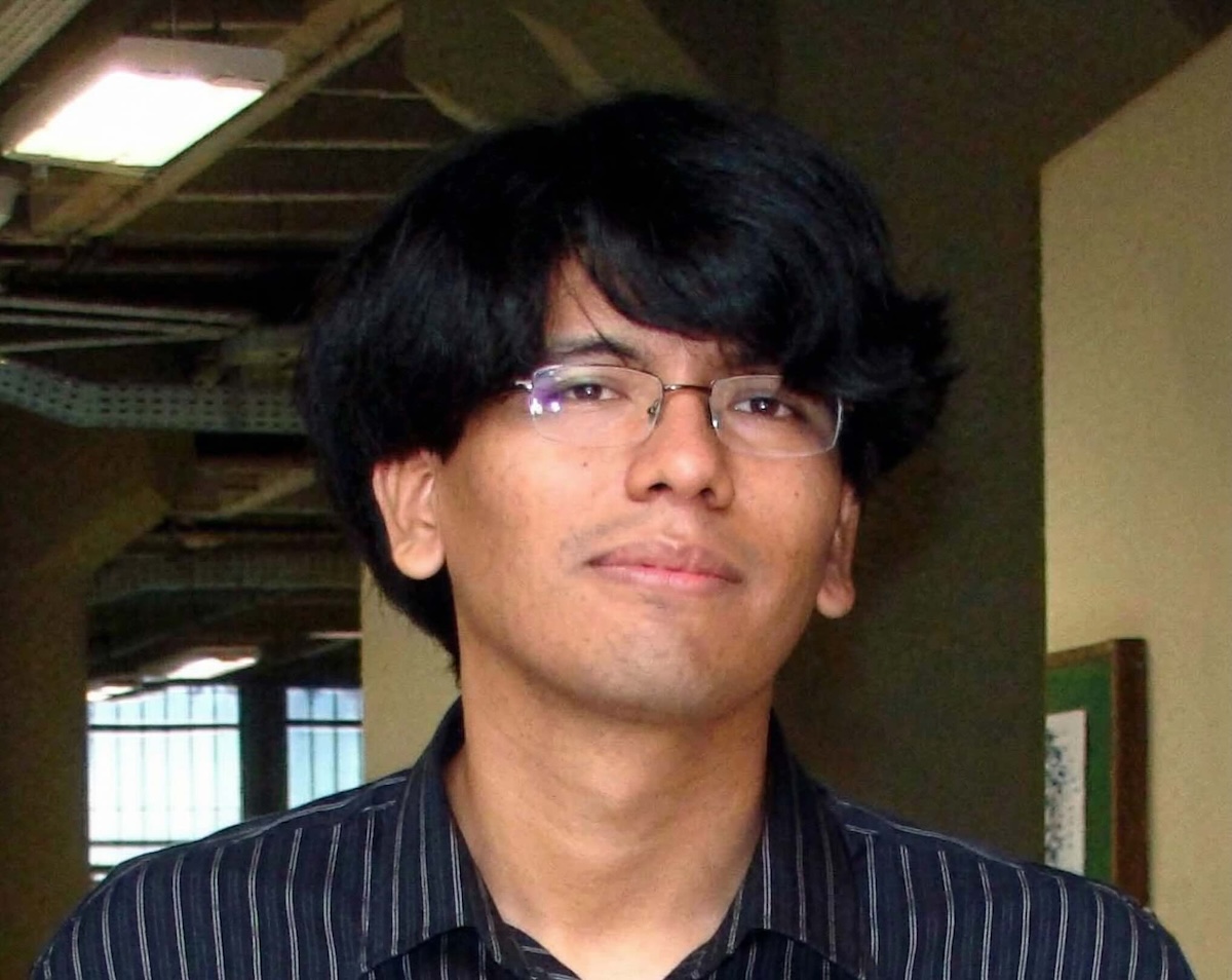 A young man with dark hair and thin-framed glasses, wearing a striped shirt and a calm expression. In the background, a corridor in an institutional setting.