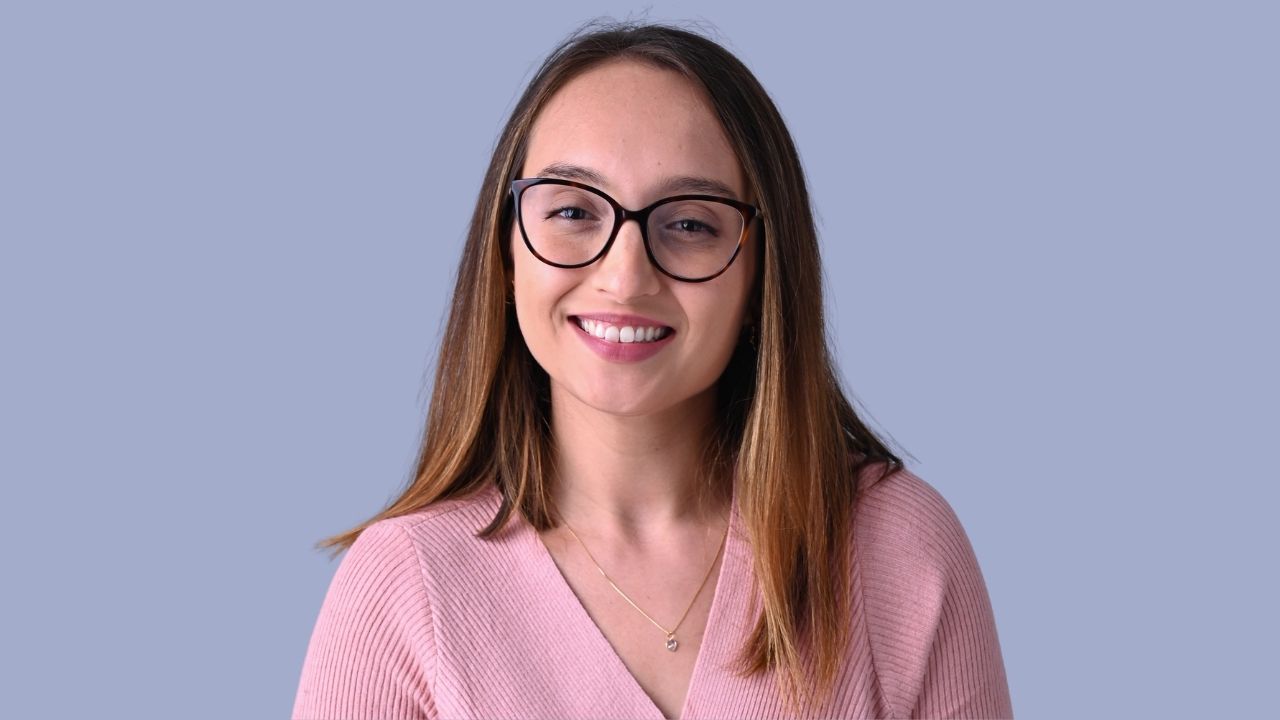 Portrait of Marina Martins Siqueira, a woman with straight brown hair with light highlights, wearing tortoise-shell glasses, a pink V-neck knit top, and a thin gold necklace with a small pendant. She is smiling and looking directly at the camera. The background is a plain bluish gray color.