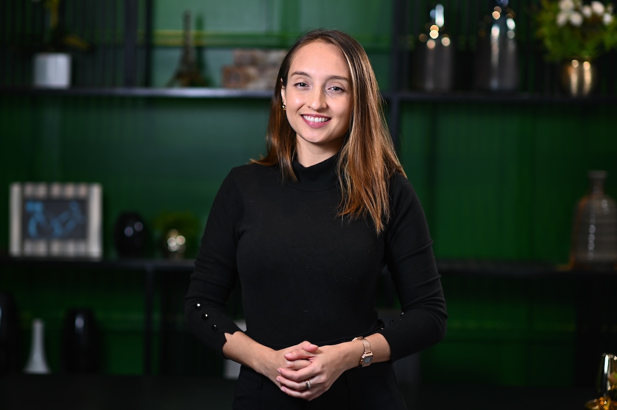 Portrait of Marina Martins Siqueira, a woman with straight brown hair with light highlights, wearing a black turtleneck sweater and a rose gold watch. With her hands clasped in front of her body, she is smiling and looking at the camera. In the background there is a dark bookcase against a green wall. The decorative objects on the bookcase are out of focus.