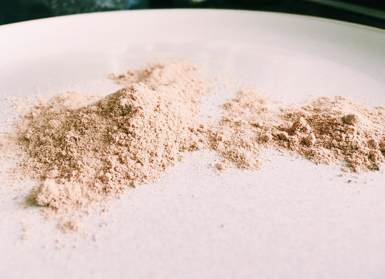 Pó de psyllium de cor bege claro espalhado sobre uma superfície branca, com textura fina e levemente granulada. A iluminação suave destaca pequenos montes e a aparência seca e fibrosa do material.
