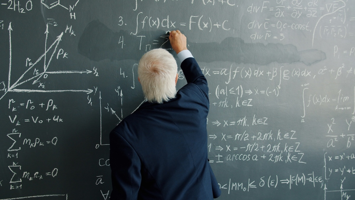 A gray-haired man, seen from behind and wearing a dark blue suit, writes mathematical formulas in chalk on a blackboard filled with equations and graphs.