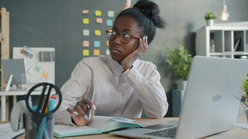 A Black woman with her hair tied back, wearing round-framed glasses, sits at her desk with a pensive expression, gazing into the distance. She rests her head on her left hand and holds a pen over an open notebook. In the foreground, there is an open laptop computer. In the background, there is a gray wall covered with colorful post-it notes, as well as plants and a bookshelf.