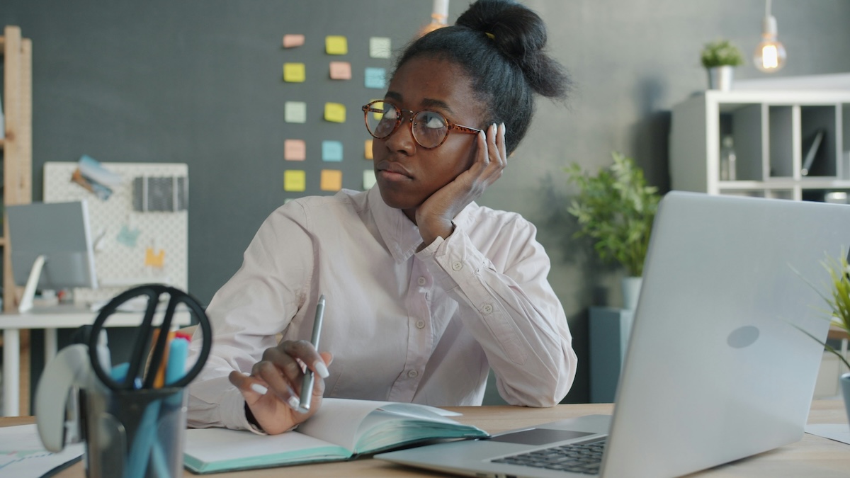 Mulher negra, de cabelos presos e óculos de armação arredondada, sentada à mesa de trabalho com expressão pensativa e olhar distante. Apoia a cabeça na mão esquerda e segura uma caneta sobre um caderno aberto. À frente, um notebook aberto. Ao fundo, parede cinza com post-its coloridos, plantas e uma estante.