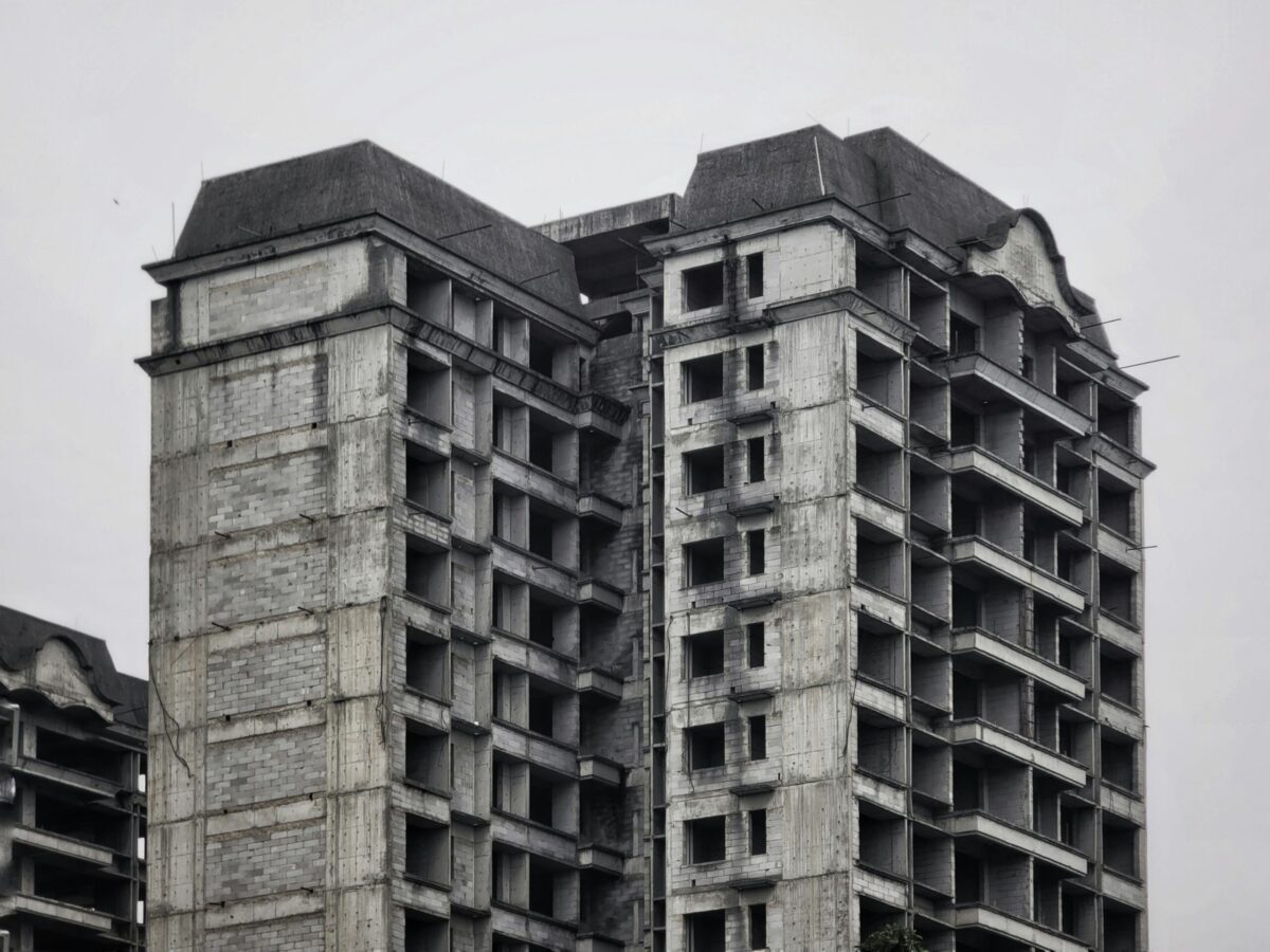 Close-up view of three abandoned high-end residential buildings, photographed in black and white under an overcast sky. The exposed concrete towers, each around fifteen stories tall, feature window openings without frames, unfinished facades, and exposed wiring.