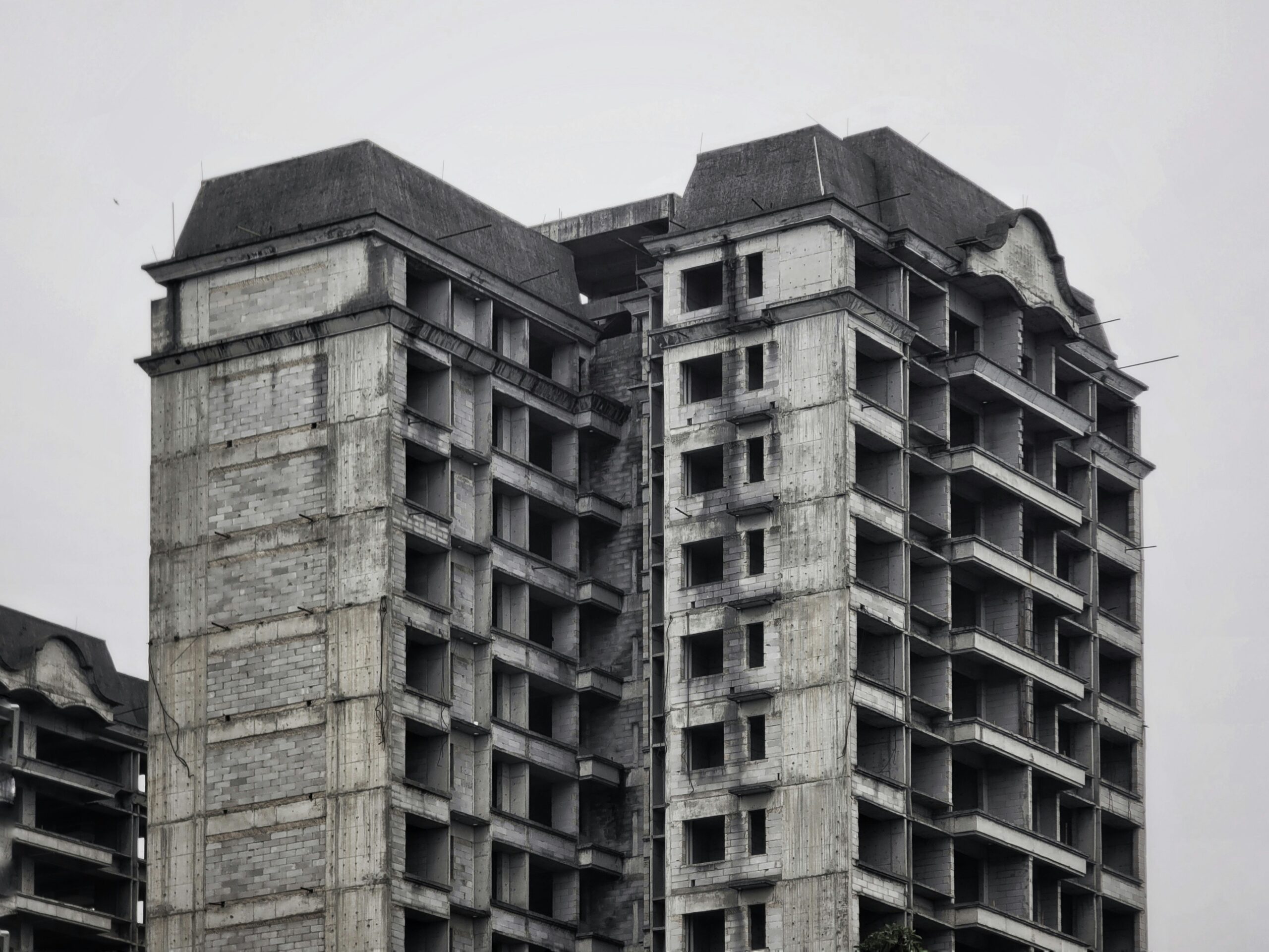 Close-up view of three abandoned high-end residential buildings, photographed in black and white under an overcast sky. The exposed concrete towers, each around fifteen stories tall, feature window openings without frames, unfinished facades, and exposed wiring.