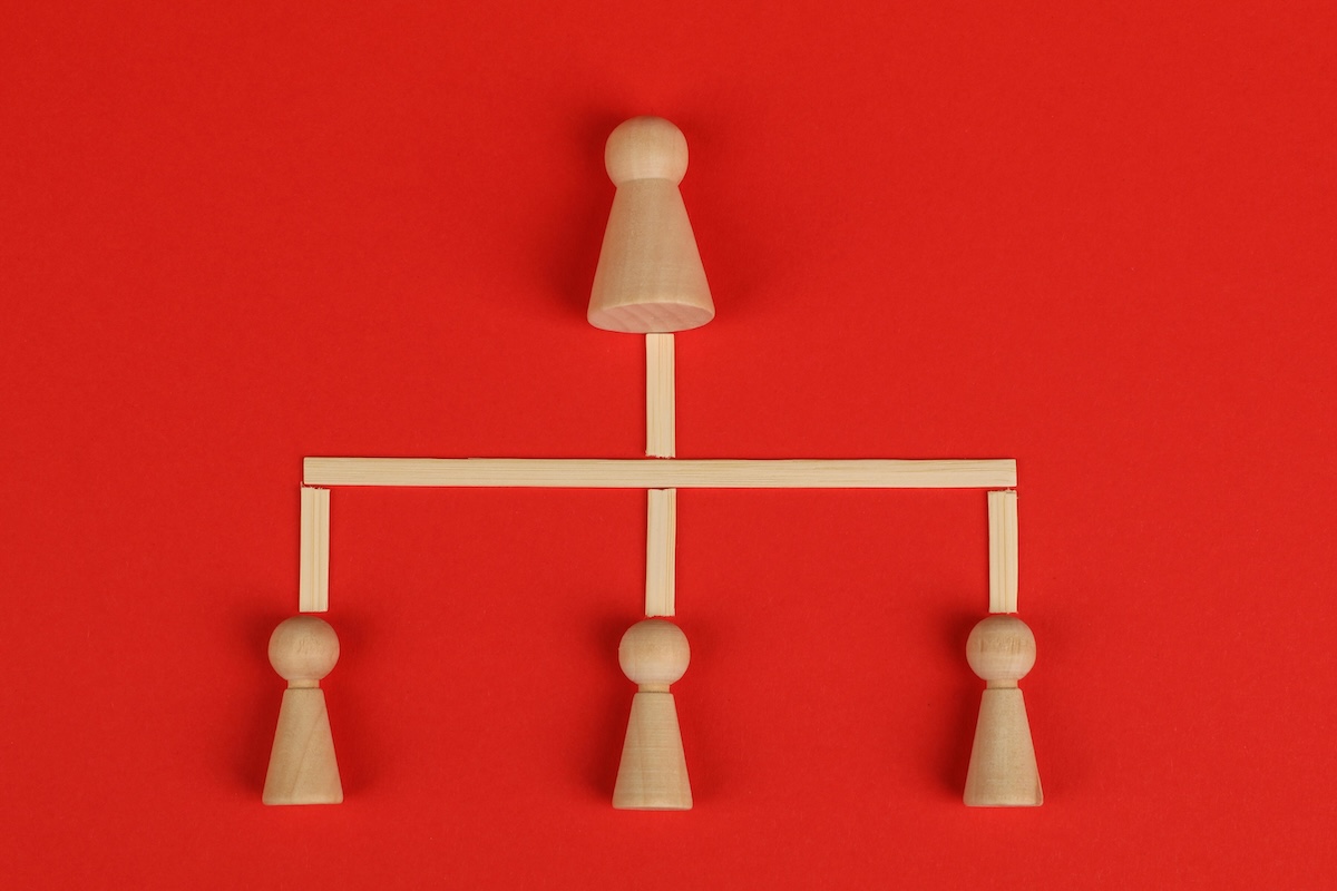 The image shows four wooden pawns arranged on a red background, forming a hierarchical structure: a larger pawn at the top, connected by wooden sticks to three smaller pawns at the bottom, representing an organizational chart.