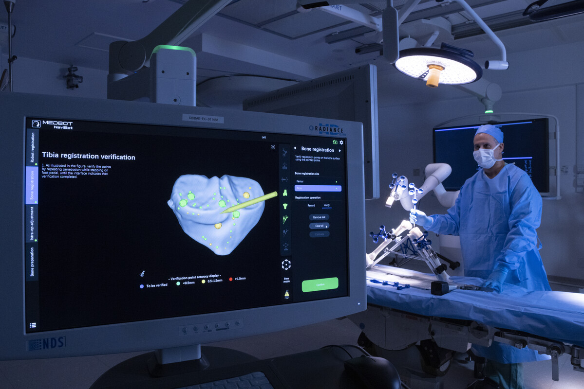 Sala de cirurgia com iluminação azulada mostra um profissional de saúde operando um sistema robótico cirúrgico, ao lado de um monitor que exibe a interface do software NaviBot, da Medbot, com modelo 3D de osso e pontos de registro para verificação de precisão em cirurgia de tíbia