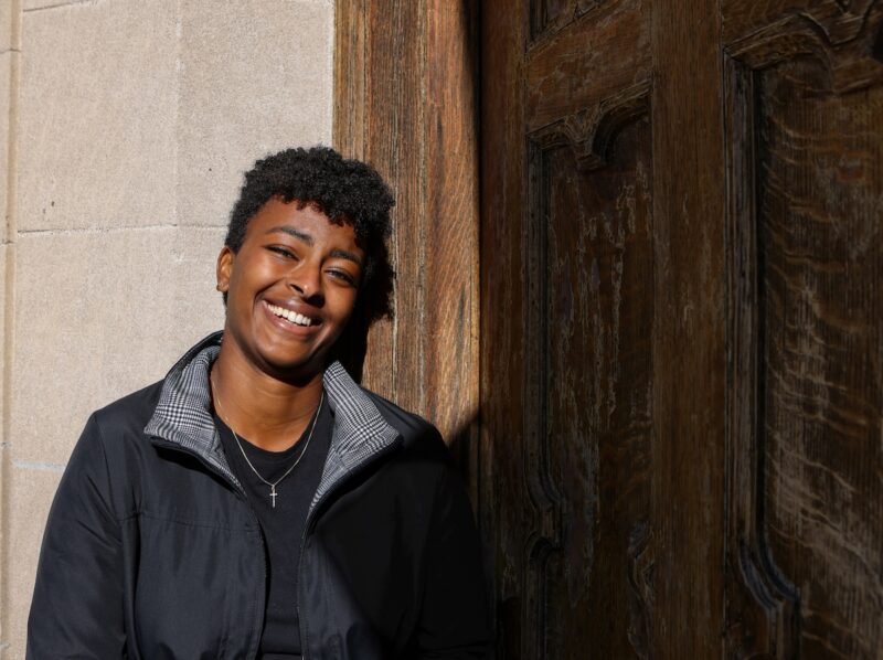 Jovem negra de cabelo cacheado curto sorrindo amplamente enquanto está encostada em uma parede de pedra ao lado de uma porta de madeira envelhecida. Ela veste jaqueta preta sobre camiseta escura e um colar com pingente em forma de cruz. A luz natural ilumina seu rosto, transmitindo bem-estar e positividade.