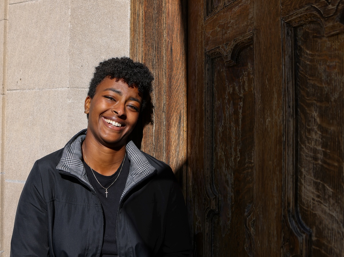 Jovem negra de cabelo cacheado curto sorrindo amplamente enquanto está encostada em uma parede de pedra ao lado de uma porta de madeira envelhecida. Ela veste jaqueta preta sobre camiseta escura e um colar com pingente em forma de cruz. A luz natural ilumina seu rosto, transmitindo bem-estar e positividade.