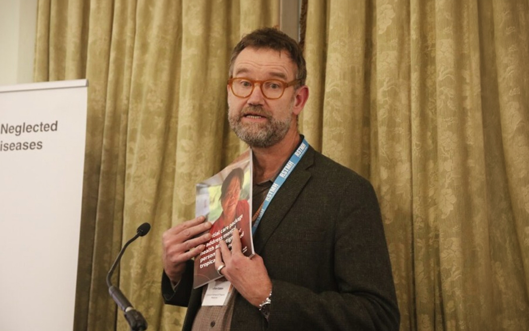 Homem branco de meia-idade, com barba grisalha curta e óculos de armação arredondada em tom âmbar, veste paletó cinza-esverdeado e usa crachá azul com a logomarca da LSTM (Liverpool School of Tropical Medicine). Está de pé, segurando uma publicação impressa com imagem de criança na capa e texto parcialmente visível que menciona "mental health" e "tropical diseases". Ao fundo, banner branco com os dizeres "Neglected Diseases" e cortinas douradas. À esquerda, suporte de microfone.