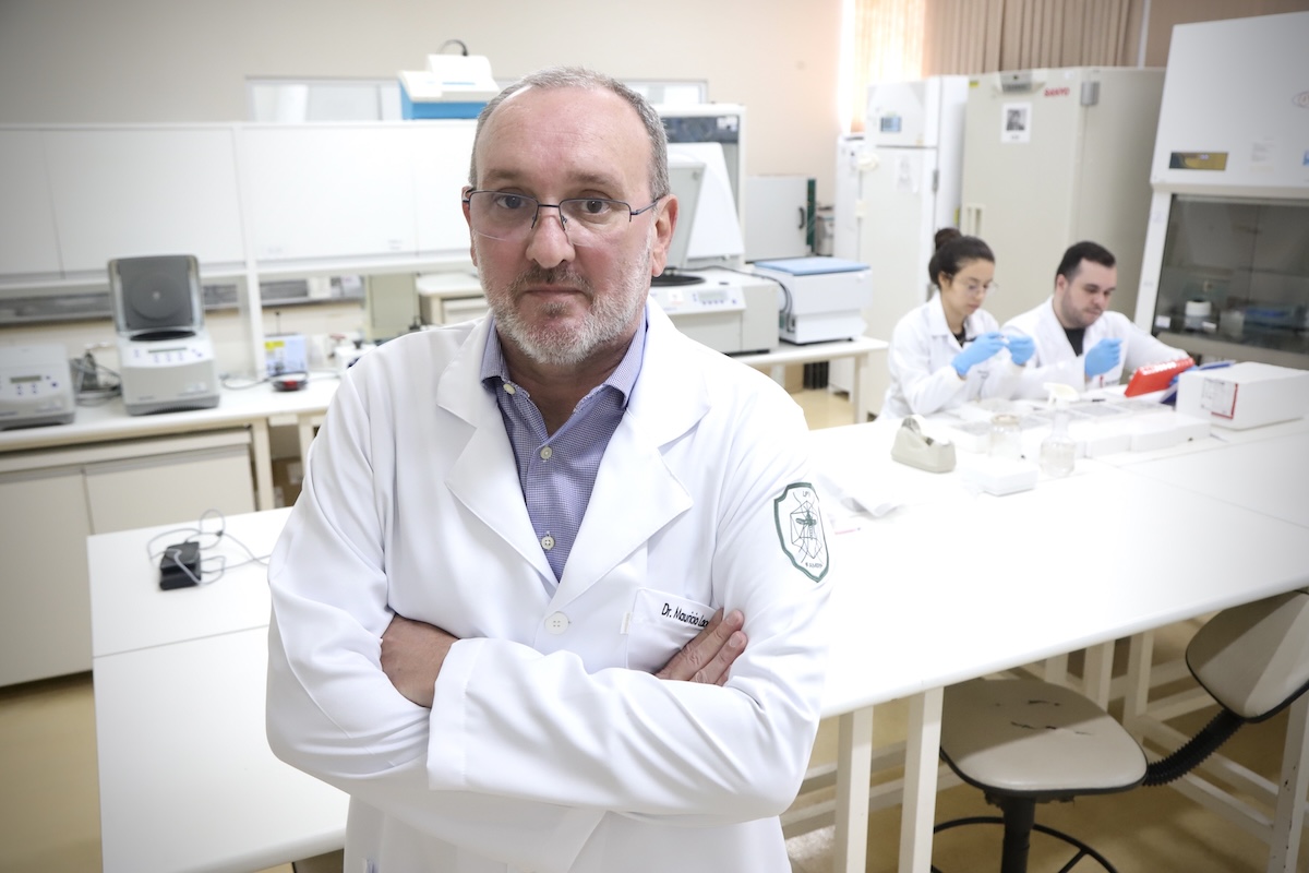 Photo of virologist Maurício Lacerda Nogueira in his laboratory at FAMERP. He is wearing a white lab coat, has his arms crossed, and is looking directly at the camera. In the background, two researchers are working with laboratory equipment.