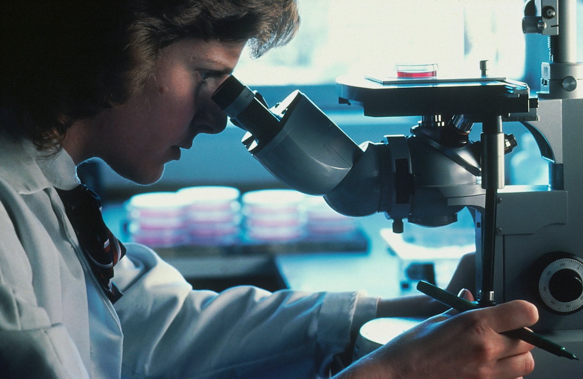 A imagem mostra uma mulher de jaleco branco e gravata utilizando um microscópio em ambiente de laboratório. Ela segura uma caneta e se inclina sobre o equipamento em posição de trabalho. Ao fundo, desfocados, são visíveis frascos de cultura empilhados e iluminação azulada característica de laboratório científico.