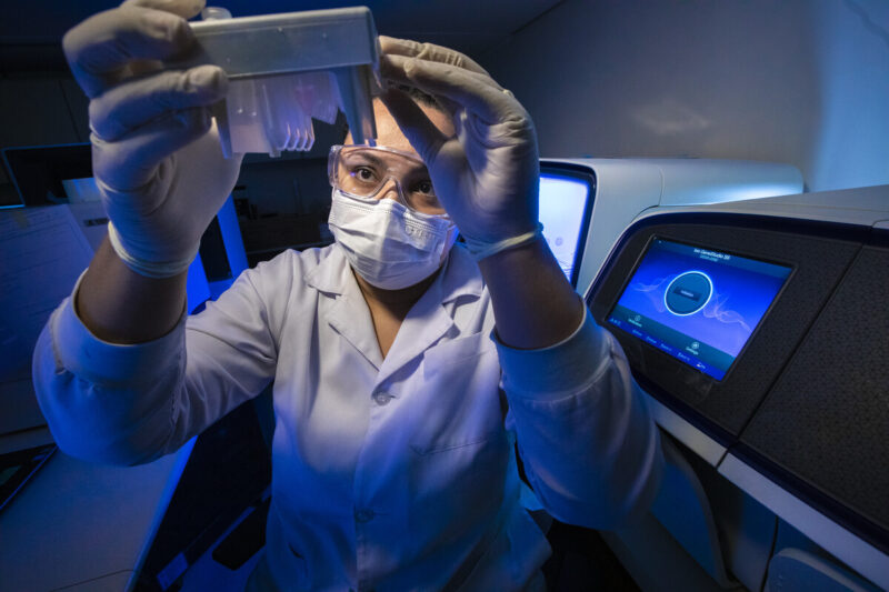 Pesquisadora com jaleco branco, máscara e óculos de proteção examina uma placa de micropoços em laboratório com iluminação azulada. Ao fundo, equipamento de sequenciamento genético com tela touchscreen ativa.