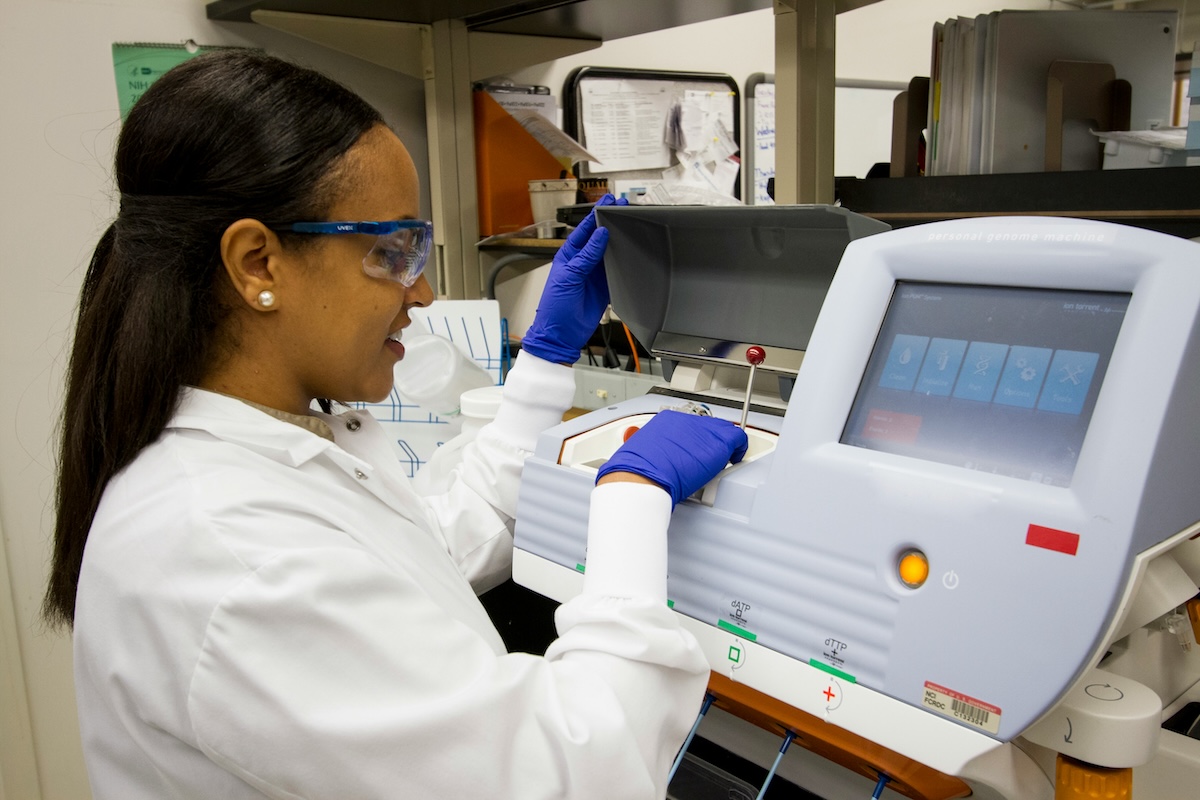 Mulher negra de jaleco branco, óculos de proteção azul e luvas roxas opera um equipamento de laboratório com tela touchscreen. Ambiente de laboratório científico com prateleiras e materiais ao fundo.