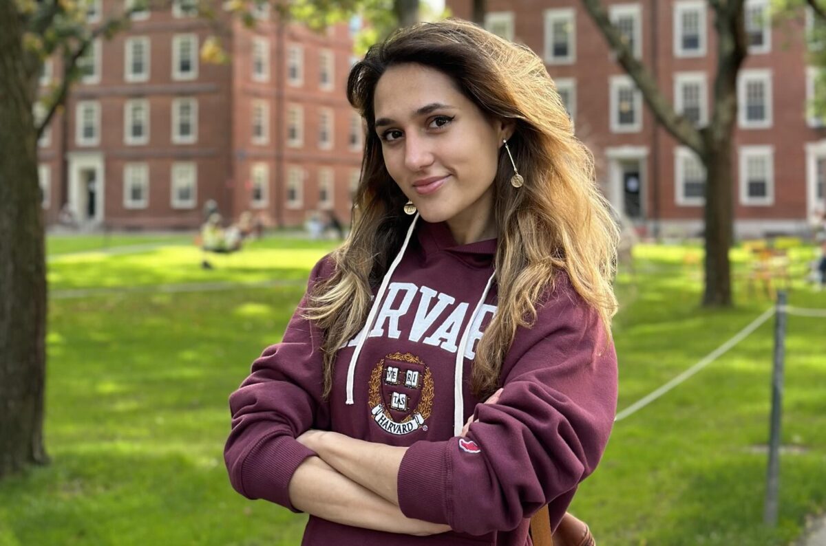 A young woman with wavy brown hair, wearing gold earrings and a burgundy sweatshirt emblazoned with the Harvard logo, stands with her arms crossed, a serene expression and a half-smile on her face, in front of a green lawn and the famous red brick buildings of the Harvard campus.
