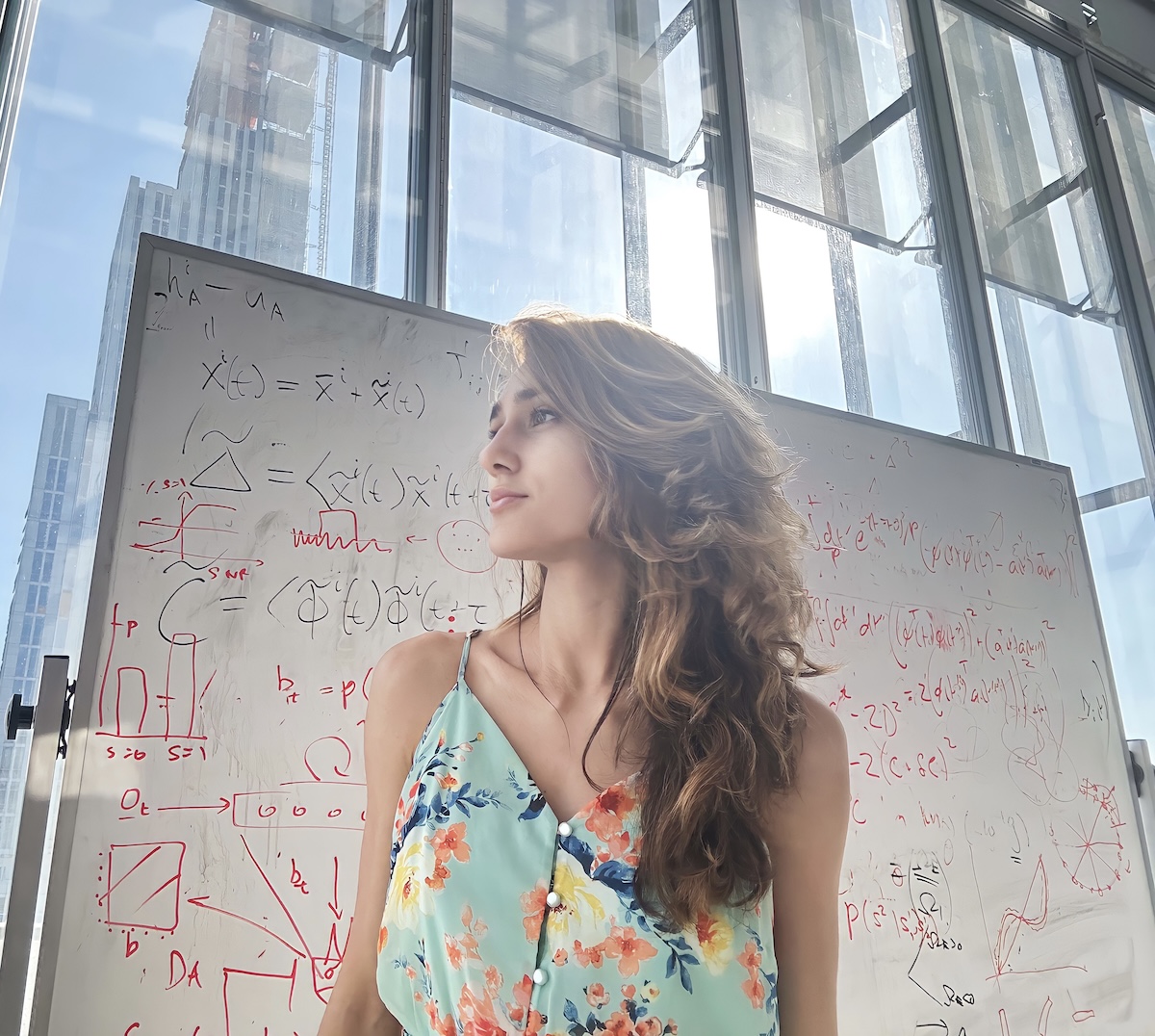 Mulher jovem de cabelos castanhos ondulados e vestido floral verde-claro, de perfil, com expressão contemplativa. Ao fundo, um quadro branco coberto de equações matemáticas escritas a vermelho e preto, e janelas amplas com arranha-céus ao fundo, sugerindo ambiente acadêmico em grande centro urbano.