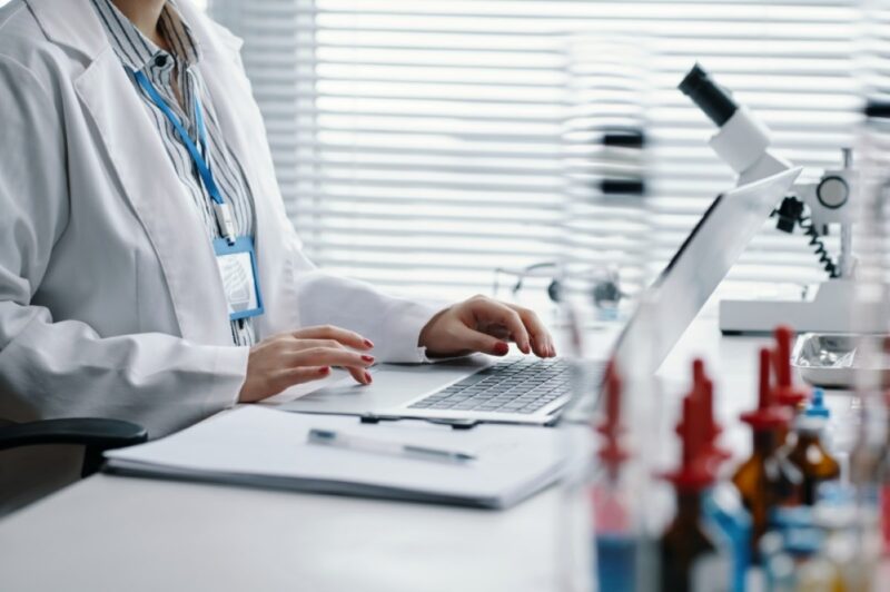 Pesquisadora em laboratório trabalha em um laptop, com microscópio e materiais científicos ao fundo