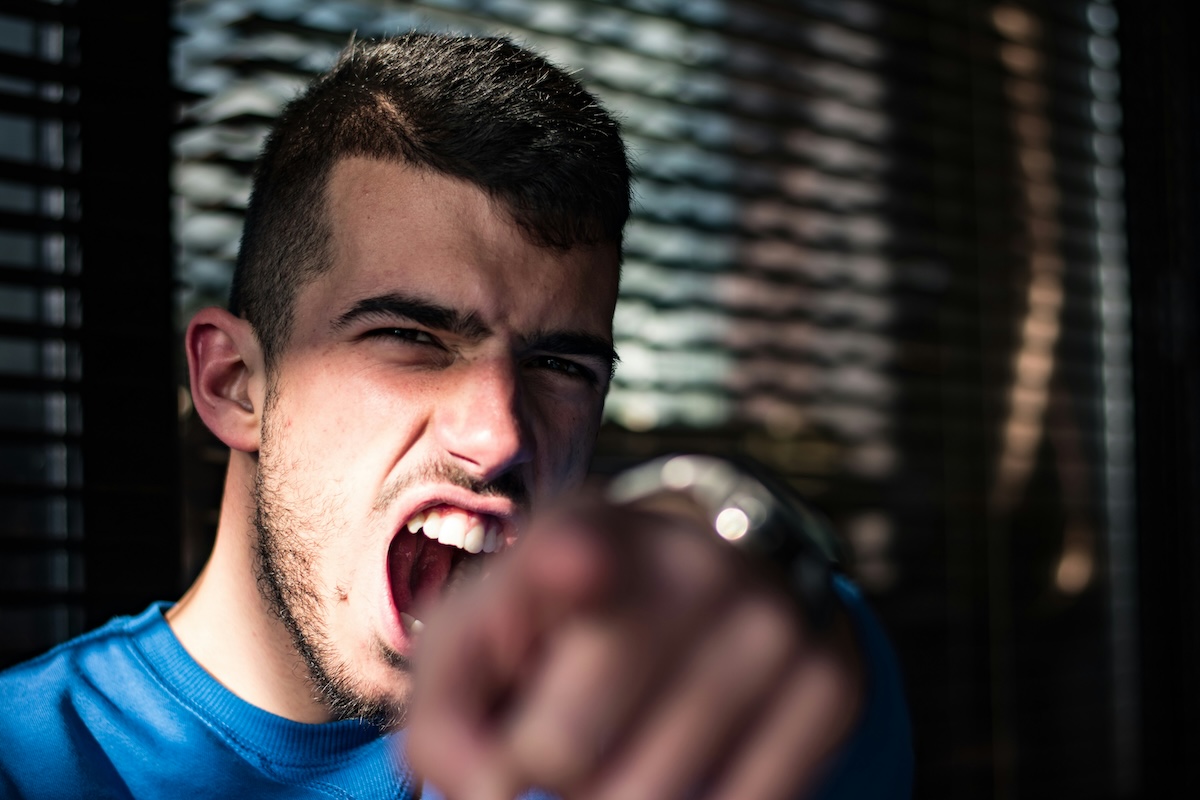 A young man wearing a blue t-shirt shouts, an expression of intense anger on his face, his fist extended toward the camera. Dark, out-of-focus blinds are visible in the background.