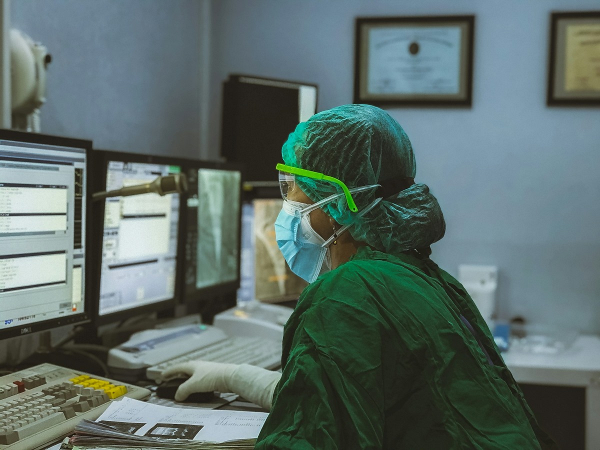 Profissional da saúde, usando máscara e roupas cirúrgicas, trabalha diante de monitores em ambiente hospitalar. A imagem remete à integração entre prática clínica, tecnologia e pesquisa científica