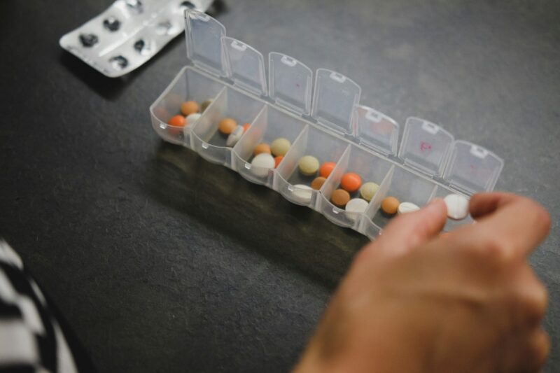 Mão retira comprimido de um organizador semanal de medicamentos, com cápsulas e pílulas coloridas distribuídas em compartimentos plásticos sobre uma superfície escura