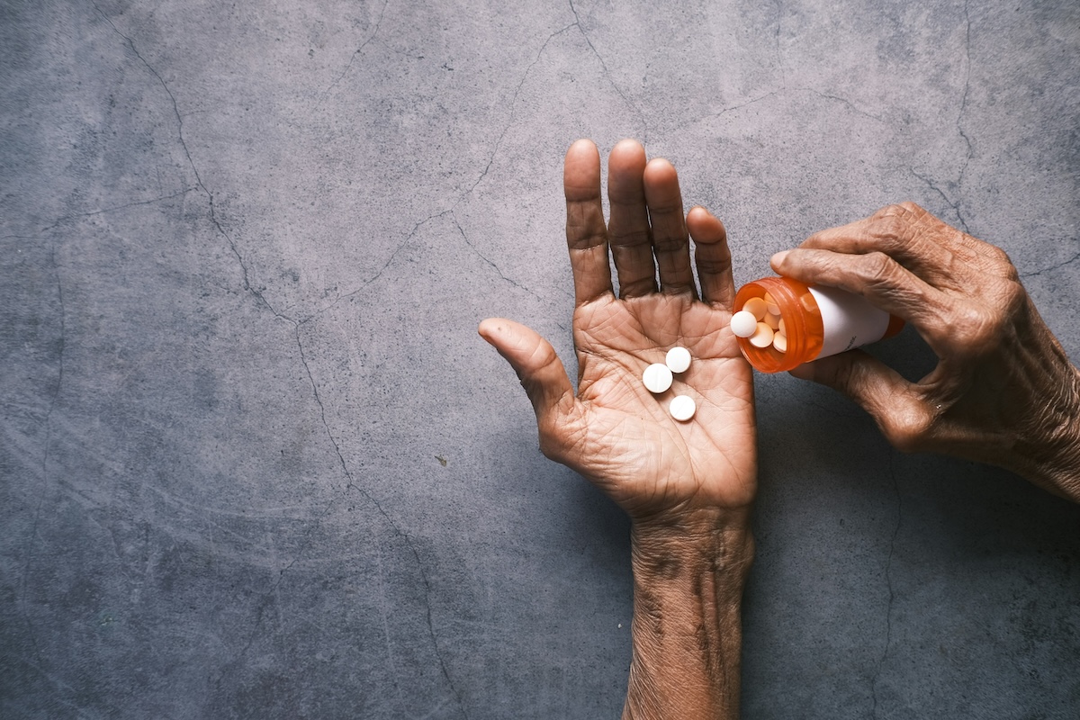 Mãos de paciente idosa seguram diferentes medicamentos sobre uma mesa, com um copo d'água ao fundo