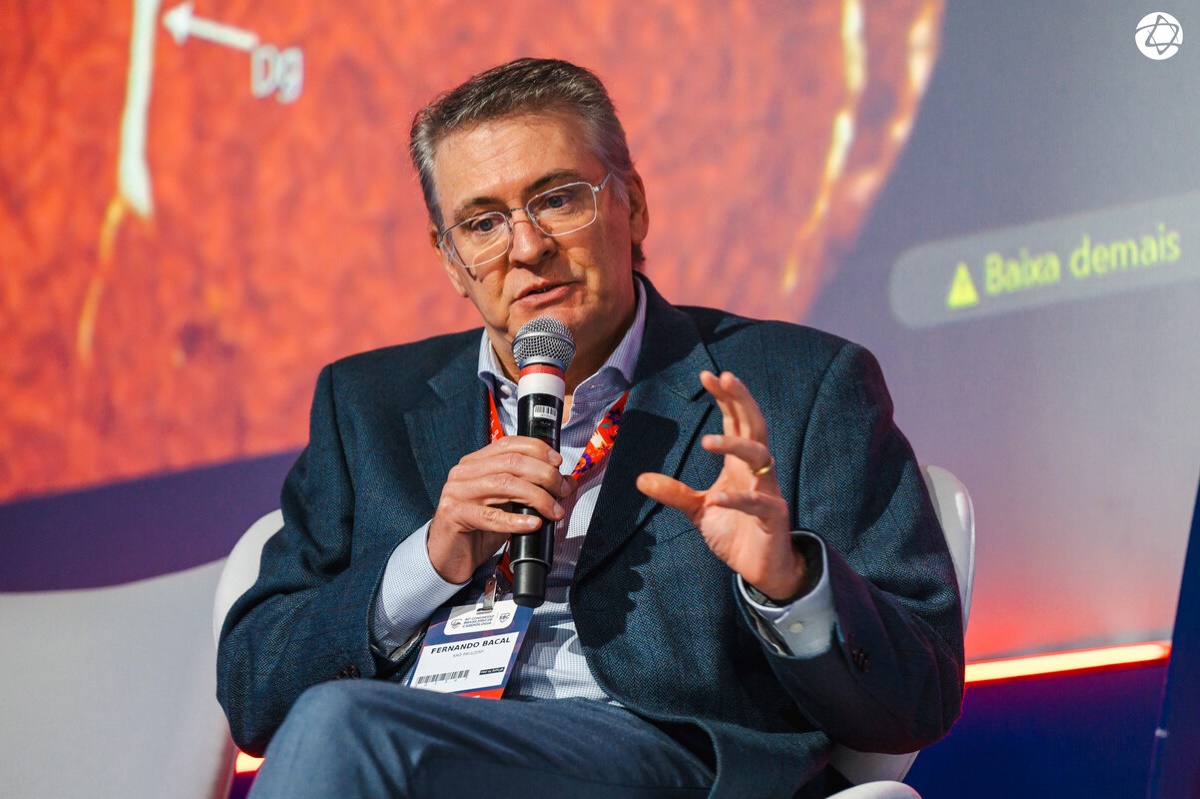 Palestrante Fernando Bacal fala ao microfone durante evento científico, com projeção ao fundo relacionada à área médica. 