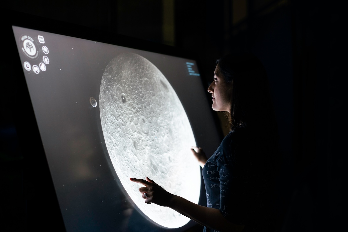 Mulher jovem de perfil em ambiente escuro aponta para tela grande e iluminada exibindo imagem detalhada da superfície da Lua; veste camiseta escura com estampas de fórmulas matemáticas