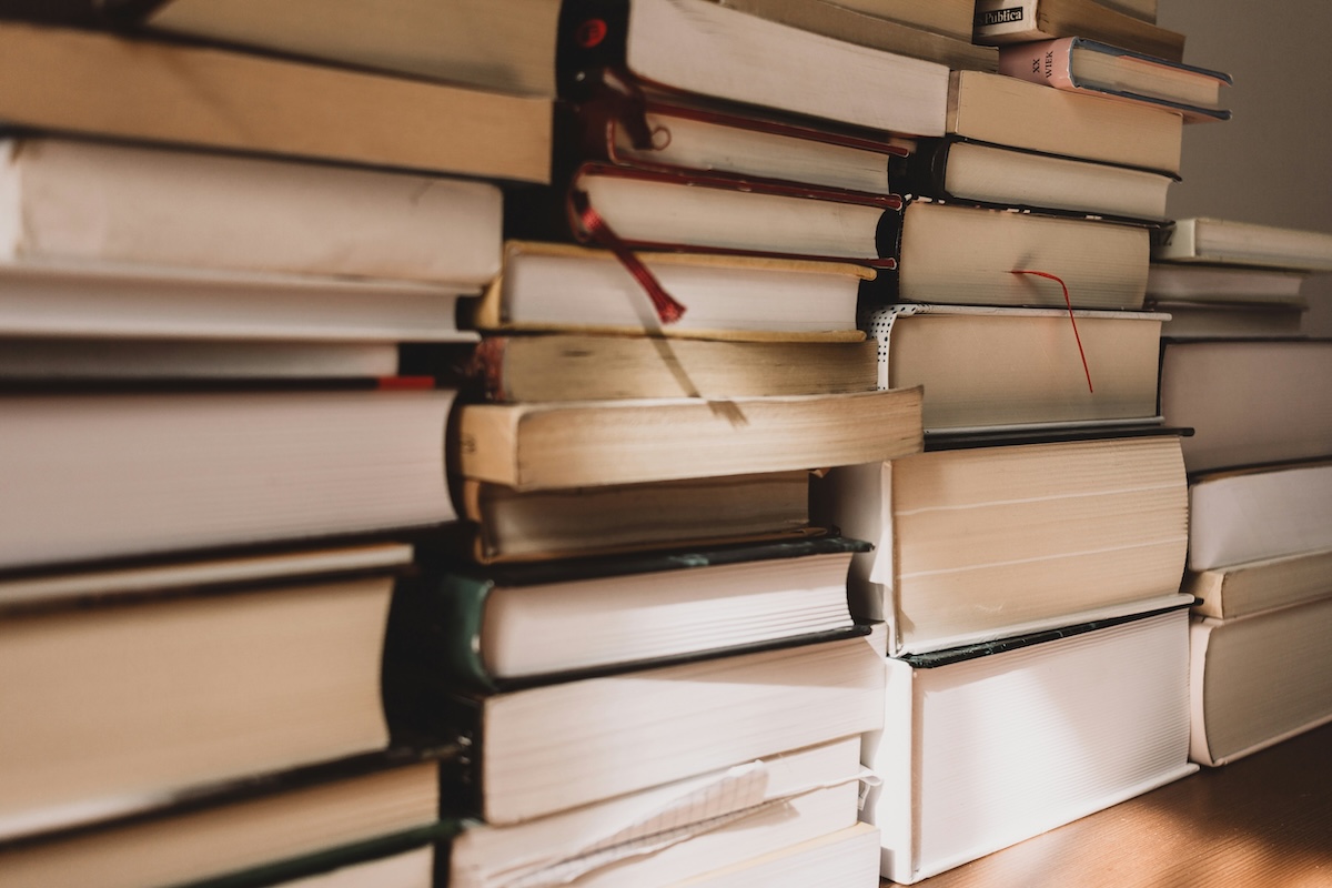 Pilhas de livros empilhados desordenadamente sobre superfície de madeira, fotografados em perspectiva com foco seletivo e luz ambiente quente
