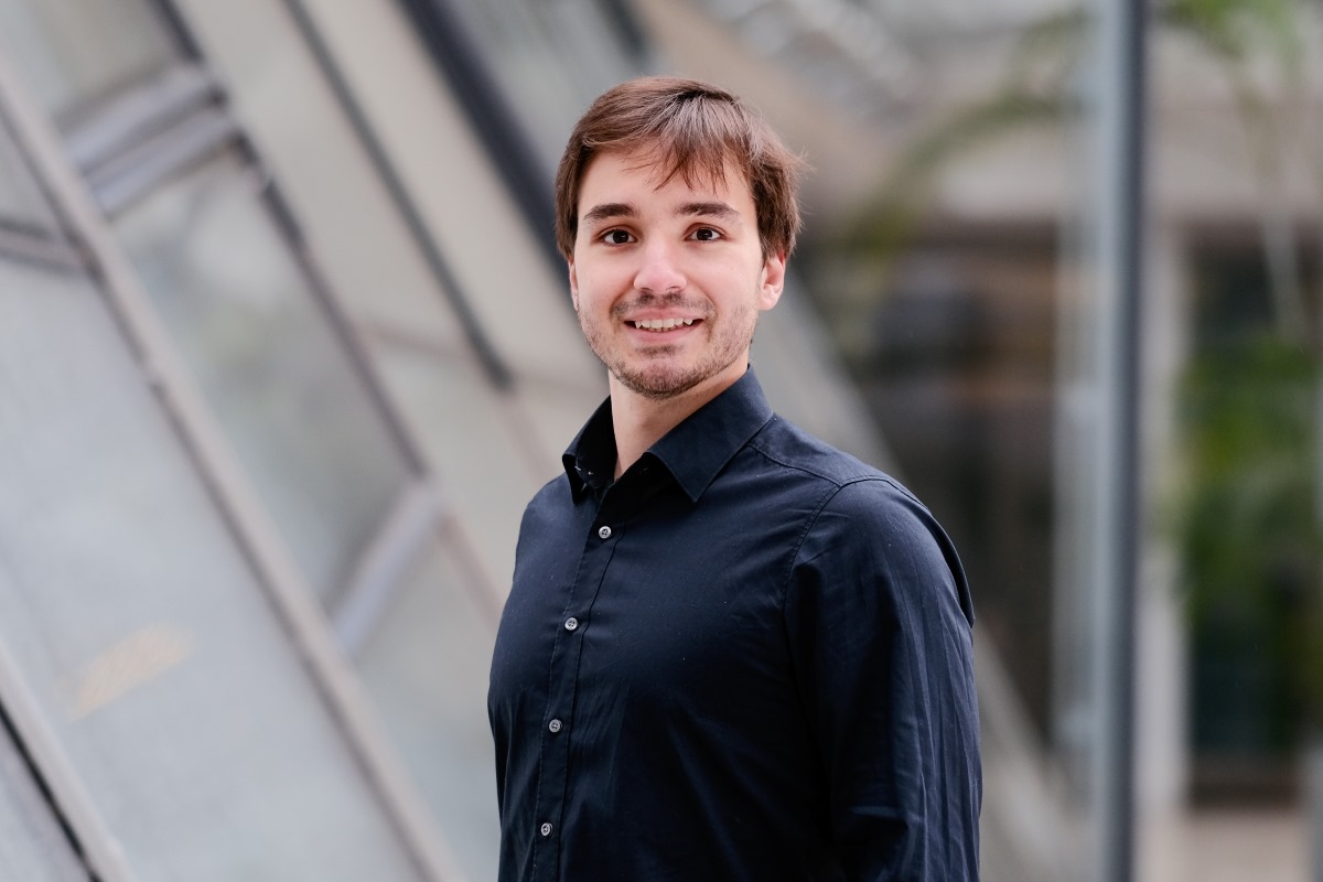 Retrato de Lennart Banse em ambiente externo, com fundo arquitetônico moderno desfocado. Ele aparece sorrindo, usando camisa escura, em uma composição de aspecto profissional