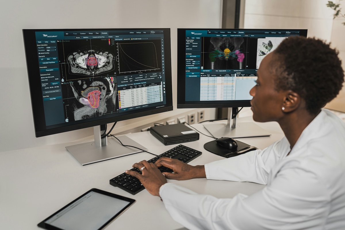 A Black female physician with short hair, wearing a white lab coat, sits in front of two monitors displaying radiotherapy planning software, with CT scan images and color-coded dose distribution maps. She has a focused expression, with her eyes fixed on the screens. On the desk there is a keyboard, mouse, and tablet.