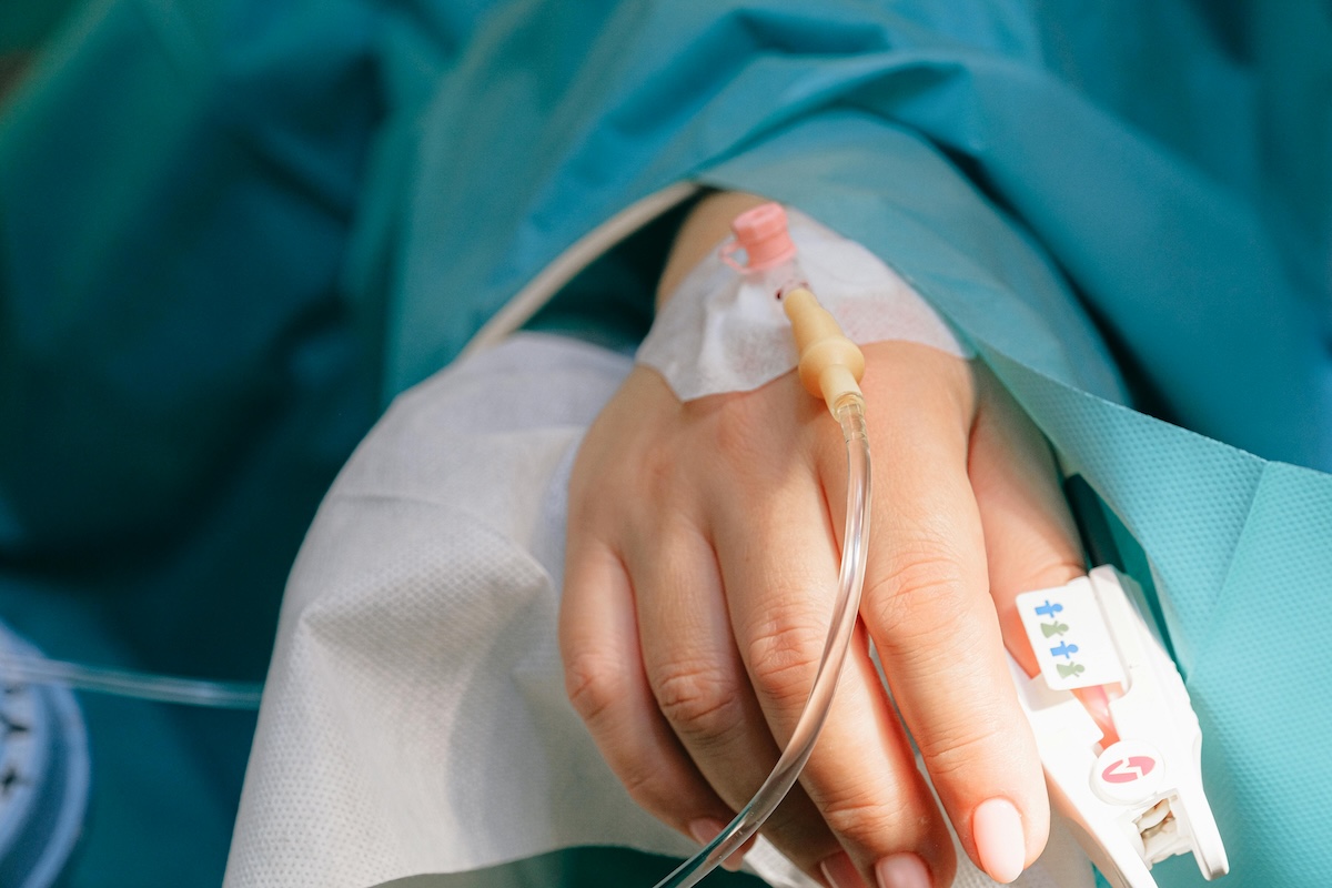 A woman's hand with pink-painted nails, wearing a green surgical gown, with an intravenous catheter secured to the back of the hand with a white dressing and connected to a transparent tube